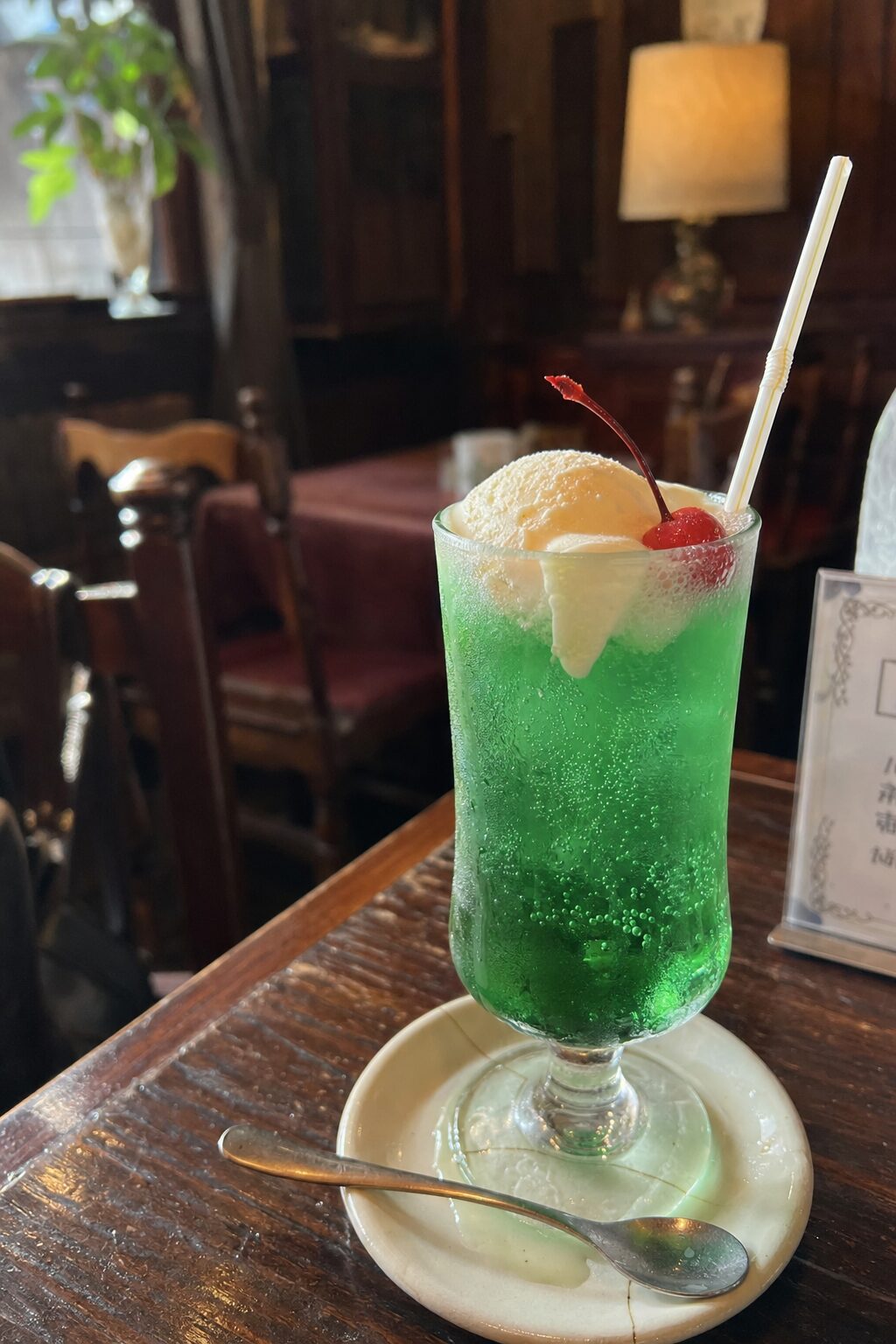 古い喫茶店の木のテーブルに置かれたメロンのクリームソーダ。緑のソーダにバニラアイスと赤いさくらんぼがのり、グラスには細かい水滴がついている。奥にランプと赤いテーブルクロスの席が見える。