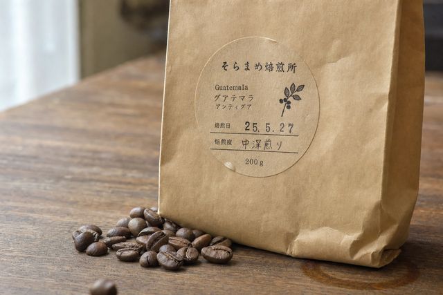クラフト紙の袋に丸いラベルが貼られ、手前にコーヒー豆がこぼれている。